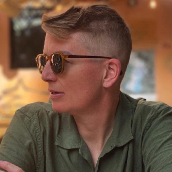 A photograph of a woman with short, close-cropped brown hair, sun glasses, and a green collared shirt who appears to be sitting in an empty resturaunt or cafe. She has her arms crossed and is looking away from the camera with her mouth slightly open.