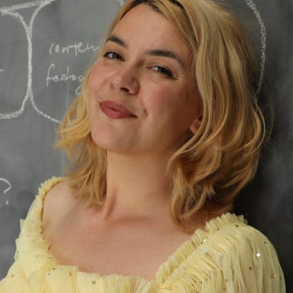 A cropped portait photo of a young woman who is leaning against a chalk board with writing on it. She has shoulder-length wavy blonde hair, dark eyebrows, red liptstick, and a yellow blouse. She is smiling and turning her head slightly toward the camera.