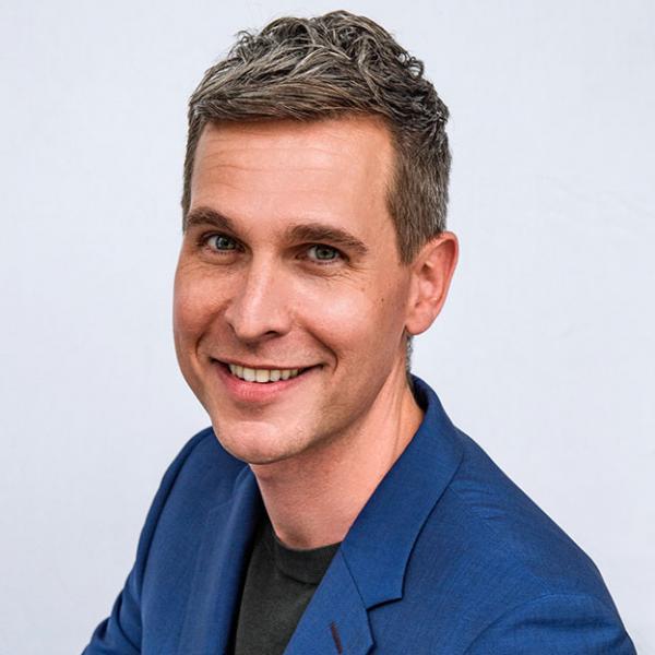 A portrait photo of a smiling man with brown hair and a royal blue blazer. He is wearing a dark grey t-shirt under the blazer and is standing in front of a blueish-white plain background.