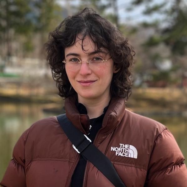 A portrait photo of a young woman who is standing outside in a park in front of a pond. She is wearing a maroon North Face puffer jacket and has the strap of a bag or purse drapped across her torso. She has long curly brown hair, wireframe glasses, and is slightly smiling at the camera.