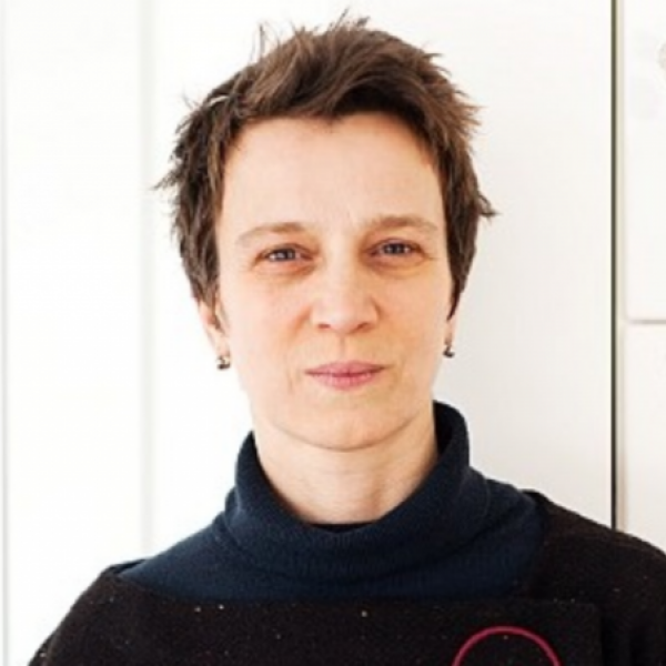 A portrait photo of a woman with short brown hair and a black turtleneck who is smiling slightly at the camera and standing in front of a white wall of cabinet doors.