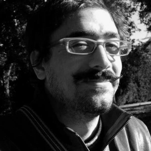 A black and white selfie photo of a smiling man with a moustache, medium-length dark hair and glasses. They are standing outside on a sunny day in what appears to be a public park.