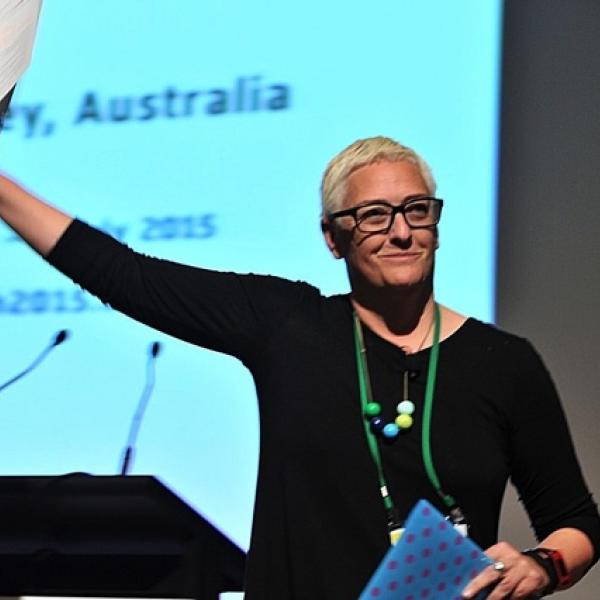 A photograph of a woman with veyr short blonder hair, black glasses, and a black shirt who is hold a piece of paper up in the air in front of a podium on a stage with a large projector on it. She is smiling slightly and looking to the right of the camera.