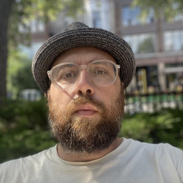 Mustafa headshot; mcu of bearded mid 40s person w/ glasses, hat, outside