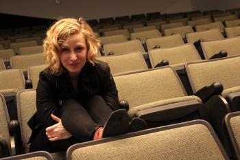 A woman with blonde medium length hair in all black is sitting alone in an auditorium.