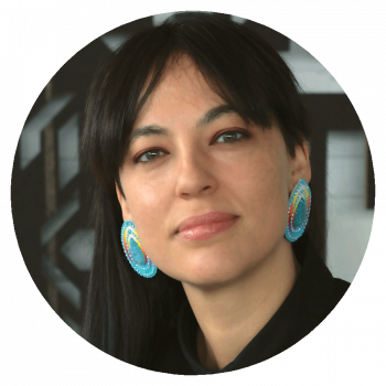 A circularly cropped photograph of a woman with black hair and colourful large earrings standing in front of a light green wall with geometric black patterns on it.