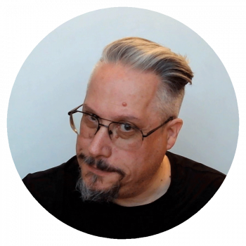 A circularly cropped portrait photograph of a man with slicked back greying hair, glasses, and a goatee. He has his head slightly tilted, is wearing a black shirt, and standing in front of a light grey wall.