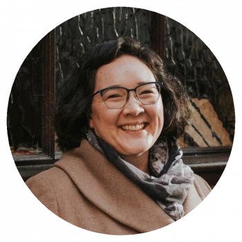 A circularly cropped portrait photograph of a smiling woman with medium-length dark brown hair, glasses, a grey scarf, and a light brown jacket who is standing in front a dark, textured wall.