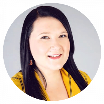 A circularly cropped portrait photograph of a smiling woman with long black hair and bright yellow clothing who is standing in front of a light grey wall.