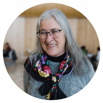 A circularly cropped portrait photograph of a smiling woman with long grey hair, glasses, a colourful scarf, and a grey knit sweater who is standing in a large light brown room with people sitting at tables in the background.