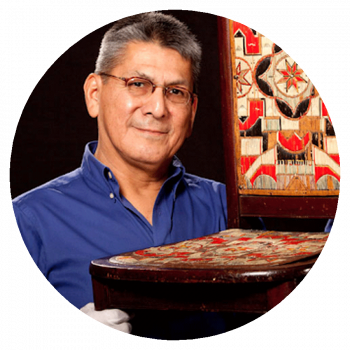 A circularly cropped portrait photograph of a man with with short greying black hair, glasses, a blue collared shirt, and white gloves who is holding an antique chair with dark stained wood and red, beige, and black inlay patterns.