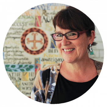 A circularly cropped portrait photograph of a smiling woman with short dark brown hair, glasses, earrings, a colourful jacket, and black shirt who is standing in front a work of art that is dense with text and patterns.