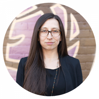 A circularly cropped portrait photograph of a smiling woman with long dark brown hair, glasses, a black coat, and a black shirt who is standing in front of an outdoor mural with brown, beige, and purple patterns.