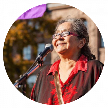 A circularly cropped photograph of a smiling woman with long grey hair, glasses, and a red shirt who is standing outside on a sunny day in front of a microphone.