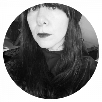 A circularly cropped black and white portrait photo of a woman with black clothing, long dark hair, and dark lipstick who is standing in an interior space.