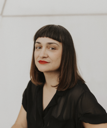 A portrait photo of a woman with long dark hair and a black blouse who is sitting in an interior space with white walls.