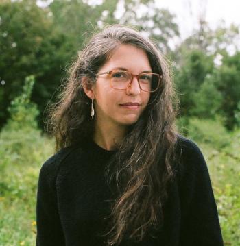 A square portait photograph of a woman with long brown hair, glasses, earrings, and a black sweatshirt who is standing in a field in front a forested area.