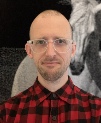 A profile photograph of a man with short dark hair, short facial hair, clear horn-rimmed glasses, and a red and black plaid-patterned shirt. He is standing in front of a wall with a large black and white photograph of a horse.