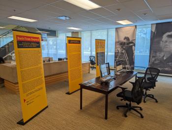 A colour photograph of a small exhibit of text and photo panels hung up around the Archives of Ontario reading room. There is also a table in the foreground with two iMacs playing archival videos, various booklets arranged on the table, and two black office chairs sitting closeby.