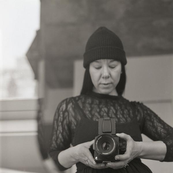A square black and white photograph of a woman with long black hair who is wearing a dark toque, a dark top, and who is holding a camera.